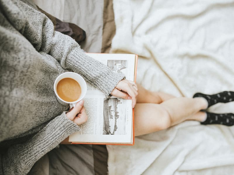 Someone resting in bed with a book and a warm drink